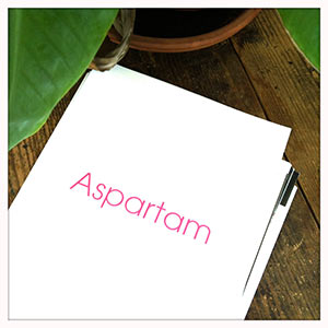 Ein Blatt Papier auf einem Holztisch auf welchem in pinker Schrift "Aspartam" steht.