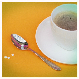Eine Tasse Kaffe auf einem orangen Tisch. Daneben liegt ein Löffel und einige Süßstofftabletten.