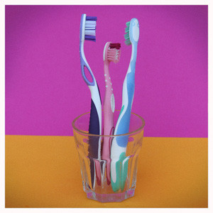 Ein Glas mit drei Zahnbürsten auf orangem Tisch mit violettem Hintergrund. Helfen Süßstoffe gesunde Zähne zu haben?