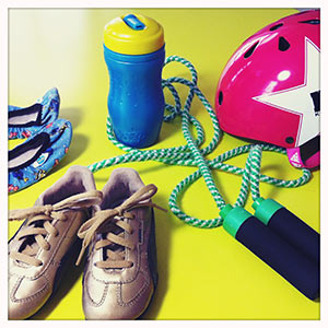 Auf grünem Untergrund stehen Sportschuhe, ein Sprungseil, eine blaue Trinkflasche und ein rosa Fahrradhelm.